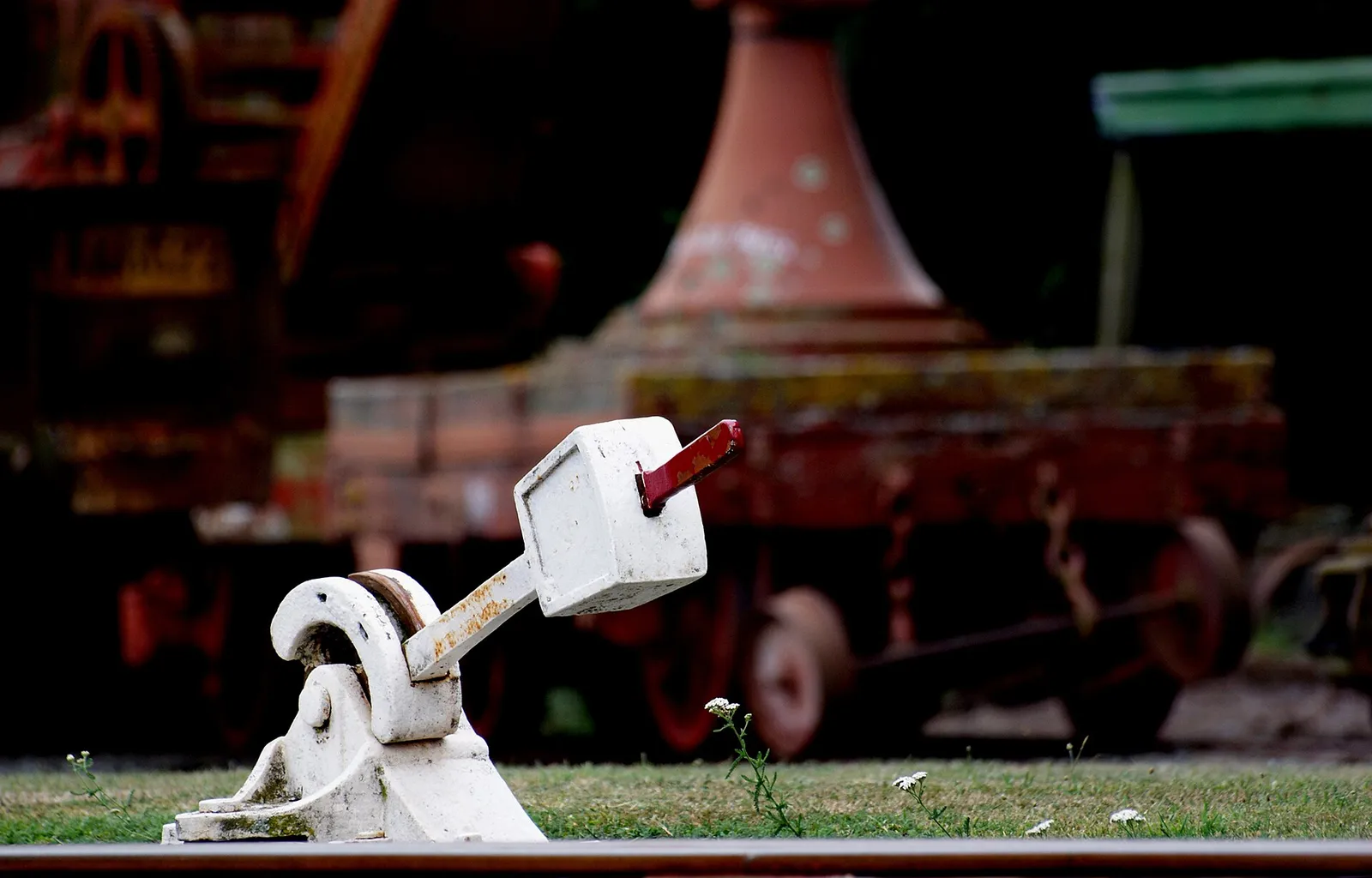 A white cast iron railway points lever with a red handle beside the rails, with a rusty old locomotive out of focus behind it