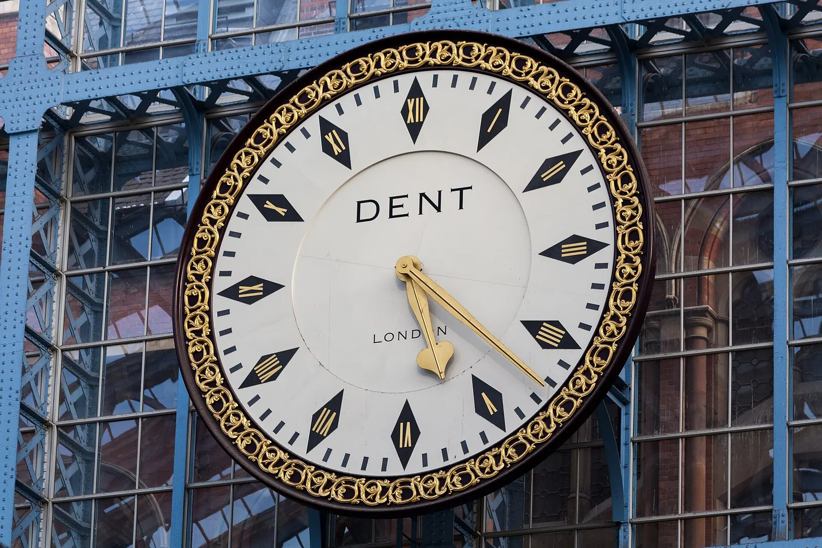 A large white station clock with black diamond hour markers and a gold scrolled rim, mounted against the blue iron and glass of a train shed
