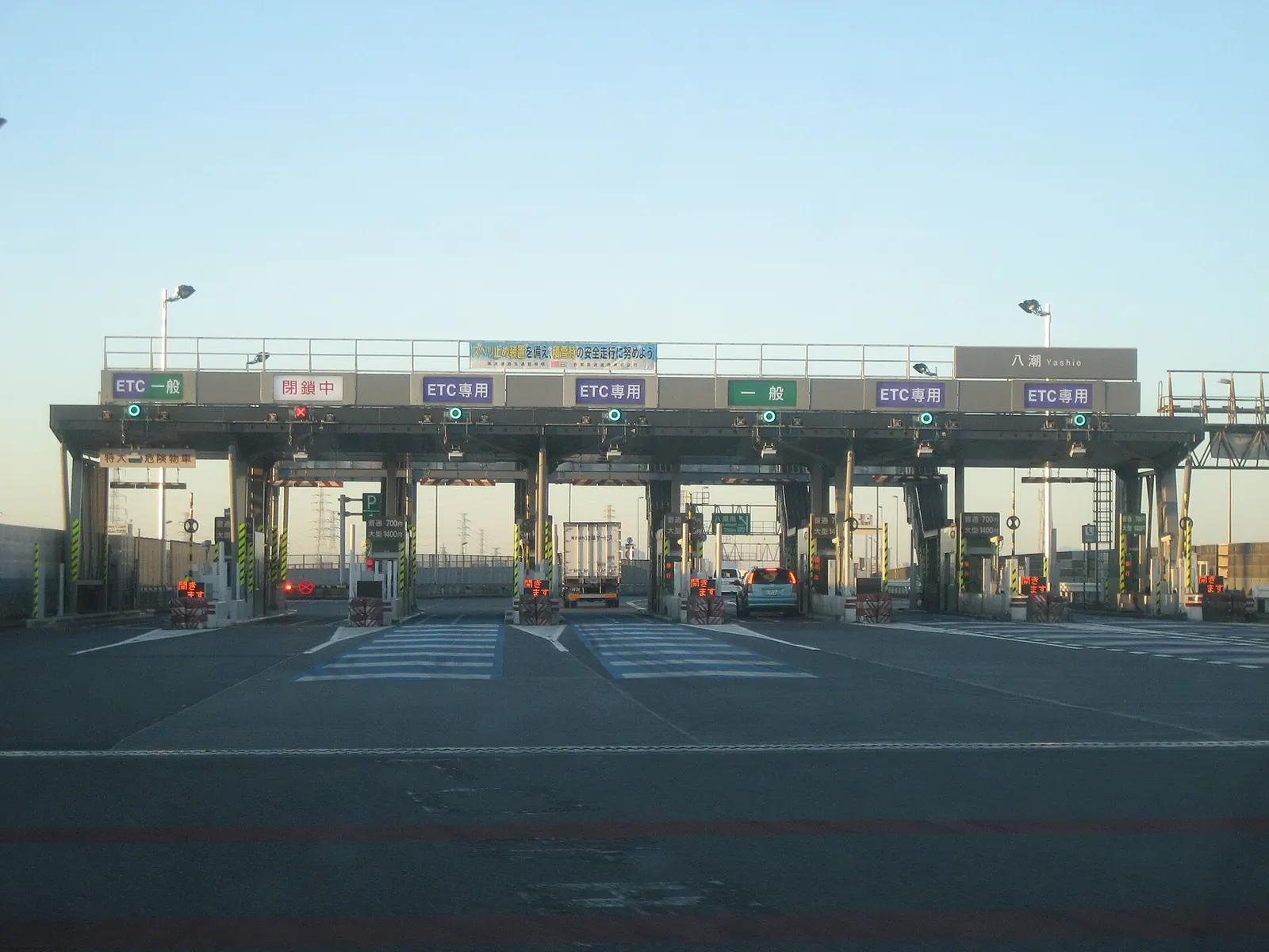 A row of highway toll booths at dawn with green and red lane signals, a few cars and a truck passing through the open lanes