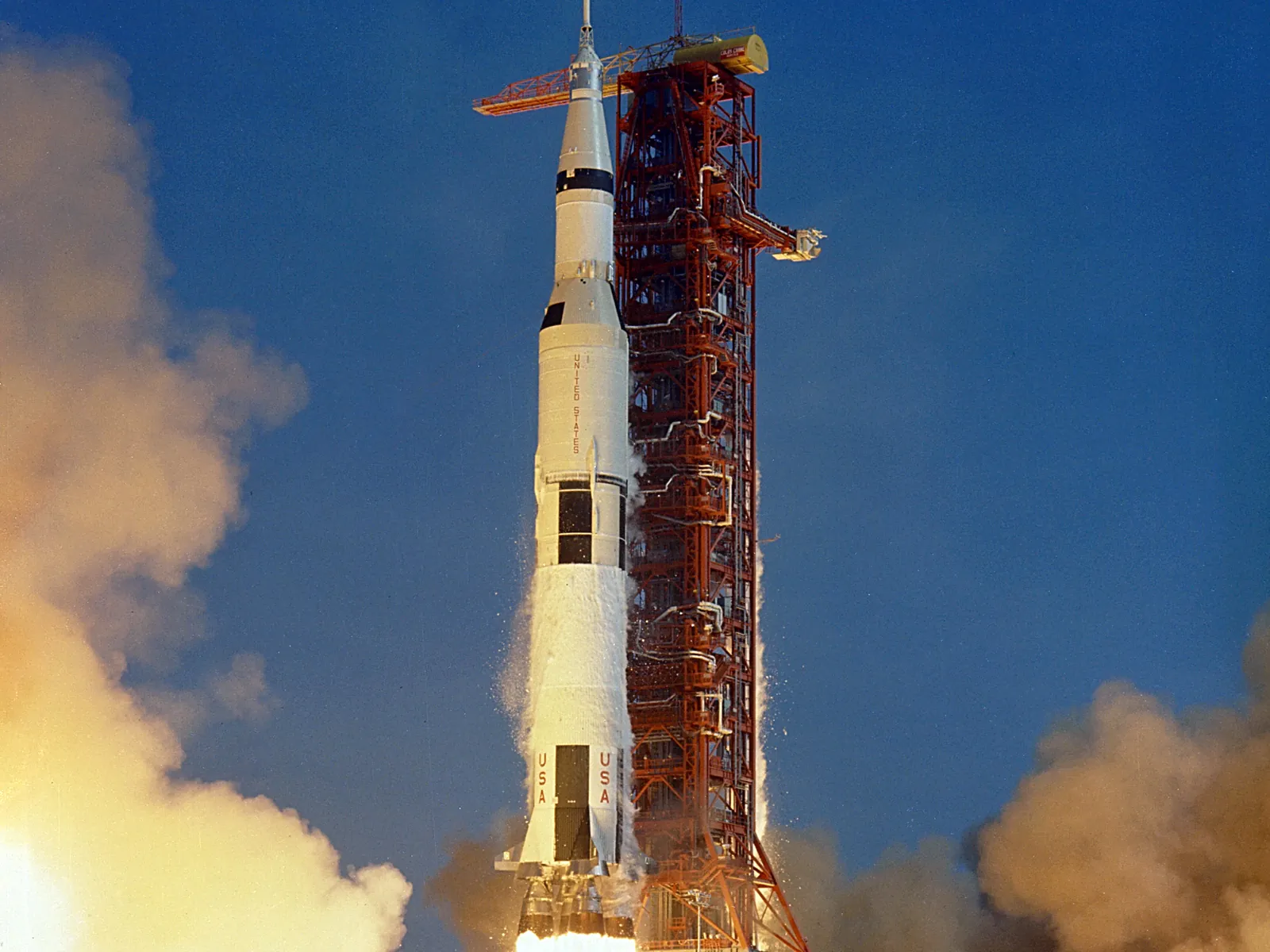 A white Saturn V rocket lifting off beside an orange launch tower, with clouds of exhaust billowing on both sides against a deep blue sky
