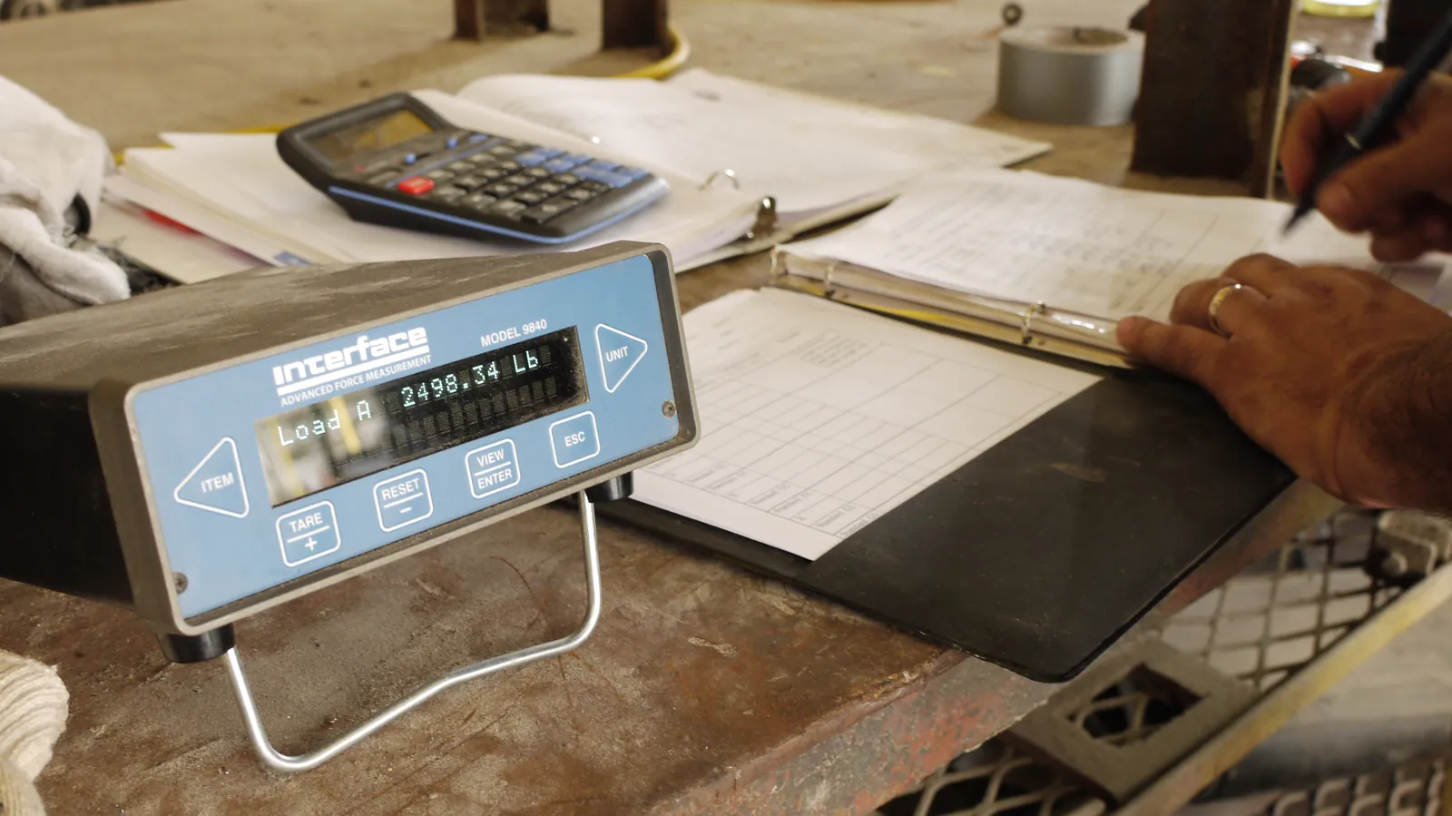 A blue digital weight indicator reading Load A 2498.34 Lb on a workbench, next to a calculator, a binder of logged readings, and a hand writing down the result