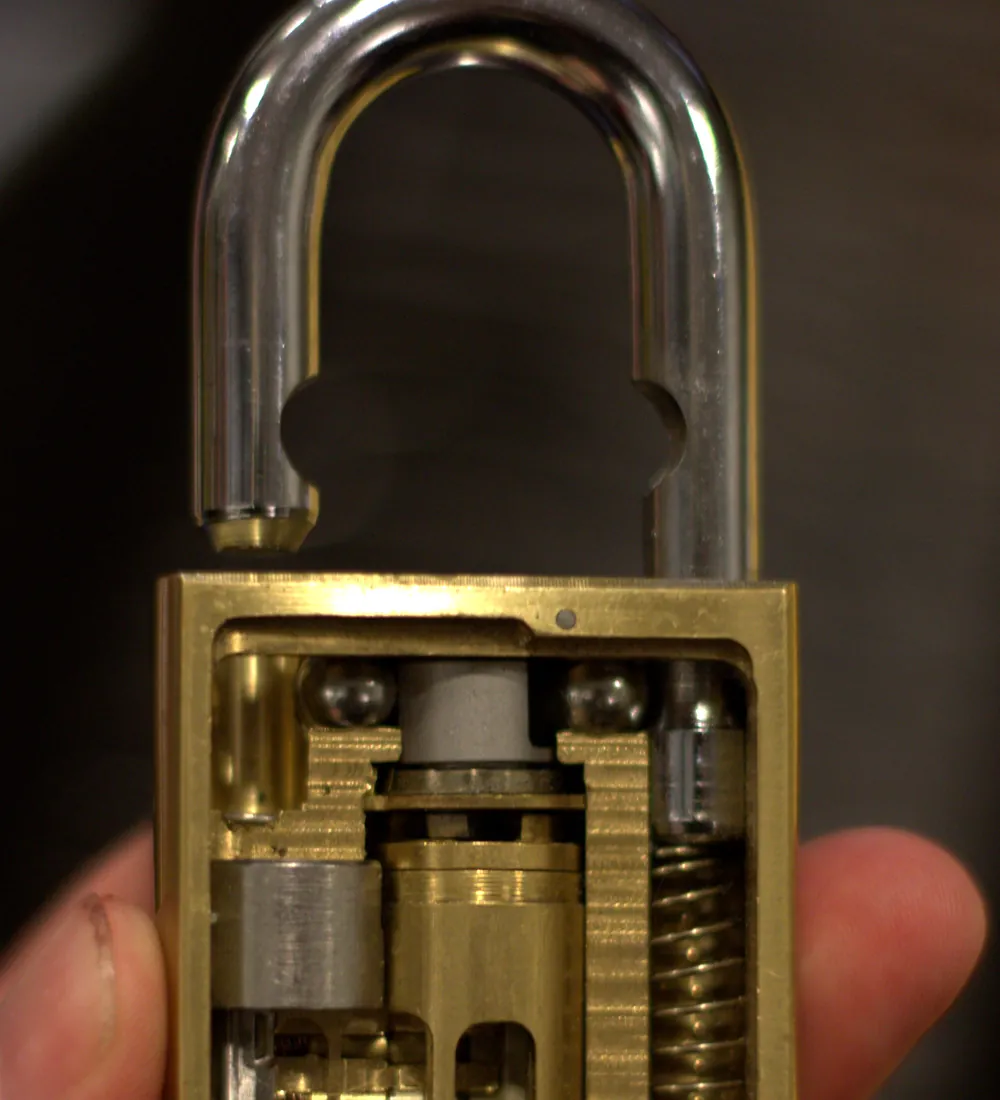 A brass cutaway padlock held open in a hand, with its shackle raised and the pins, springs, and locking balls visible through the cut-away body