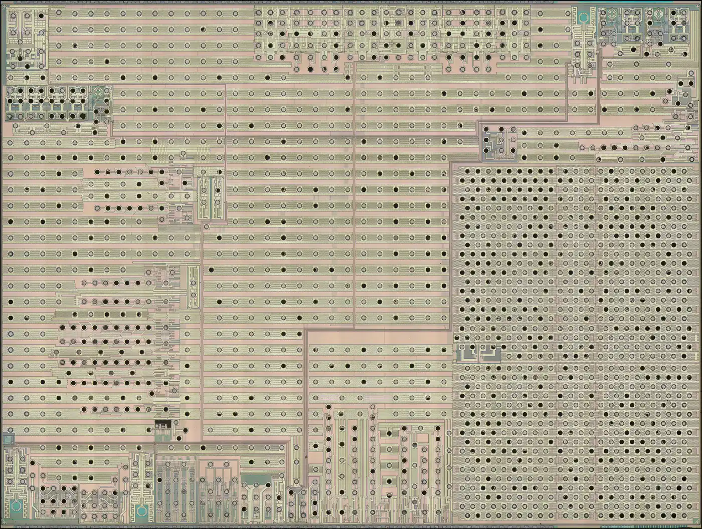 A highly magnified photo of a bare silicon die: rows of horizontal metal power rails across most of the chip, a dense grid of dark contact dots filling the right third, and small blocks of logic along the edges