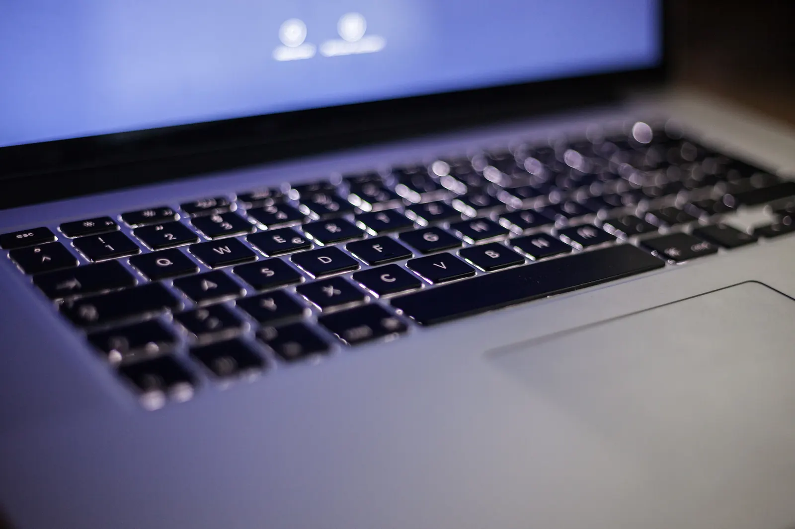 A close-up of a silver laptop’s backlit keyboard in a dark room, with the screen glowing blue at the top and the trackpad in the lower right, shot with a shallow depth of field