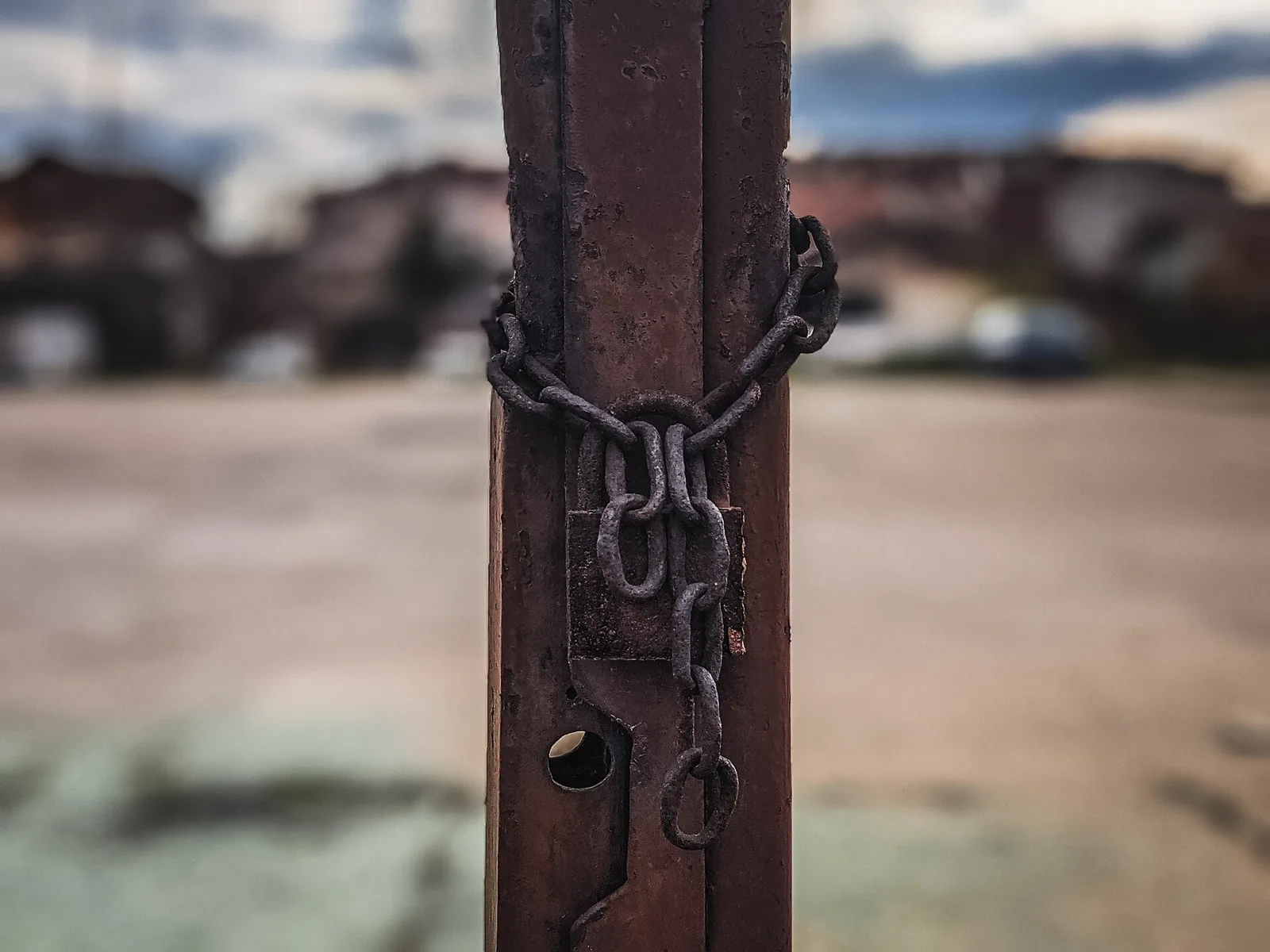 A rusty chain wrapped around a rusty steel gate post and hooked over its latch, with a blurred parking lot and houses behind it