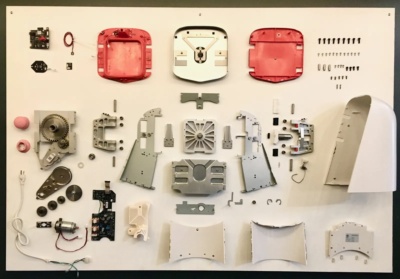A consumer kitchen appliance taken completely apart and mounted on a white board: red and white plastic housings, aluminum frames, gears, a motor, two circuit boards, a power cord, and rows of screws laid out in a grid