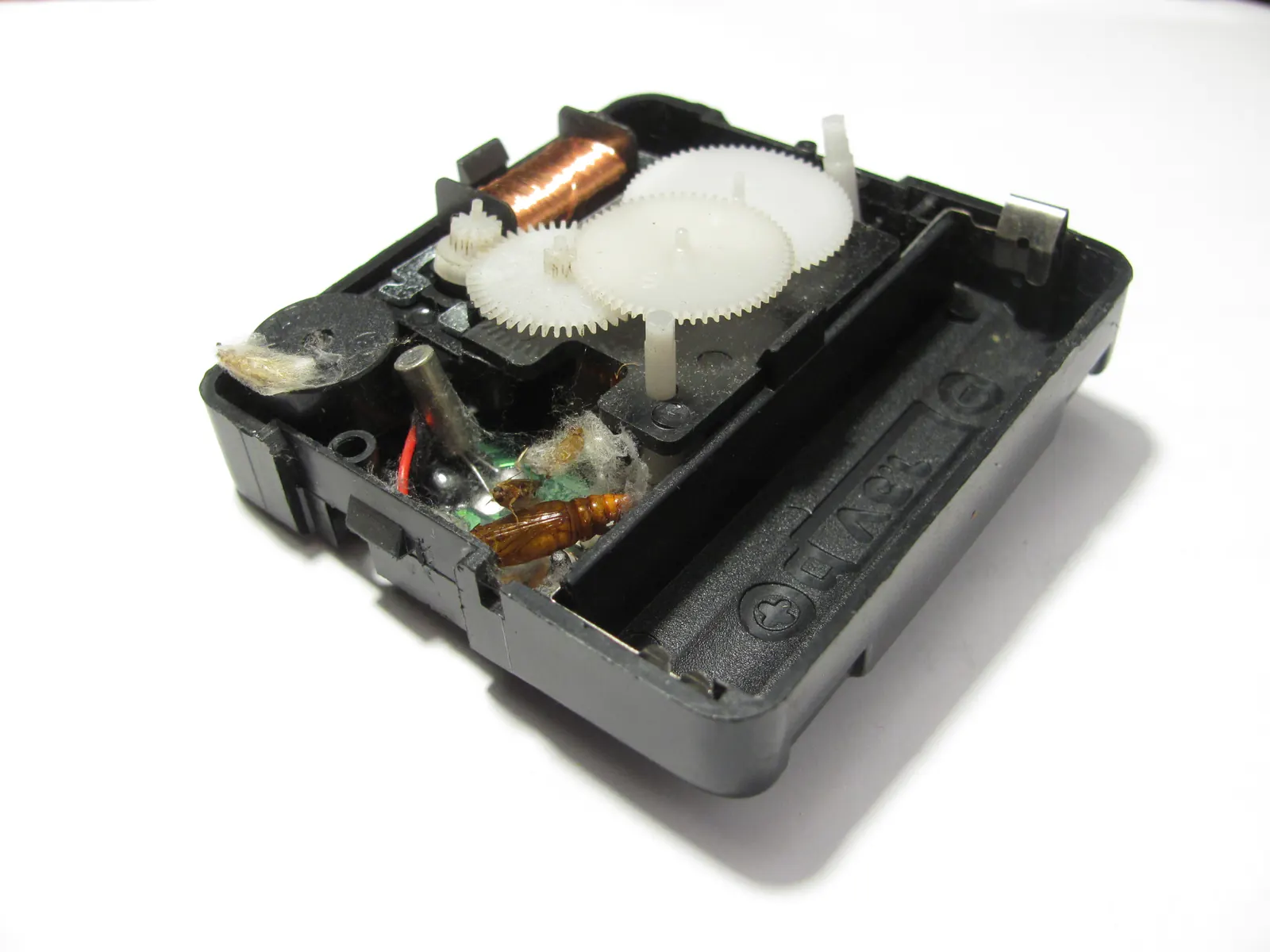 The inside of a small quartz clock movement: white plastic gears, a copper coil, a green circuit board, and a silver button cell, all exposed